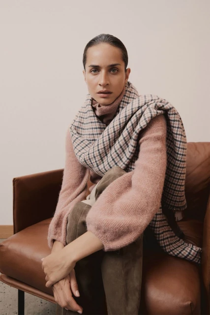 A stylish adult female sits indoors on a brown leather chair wearing a pink sweater and plaid scarf, looking directly at the camera.