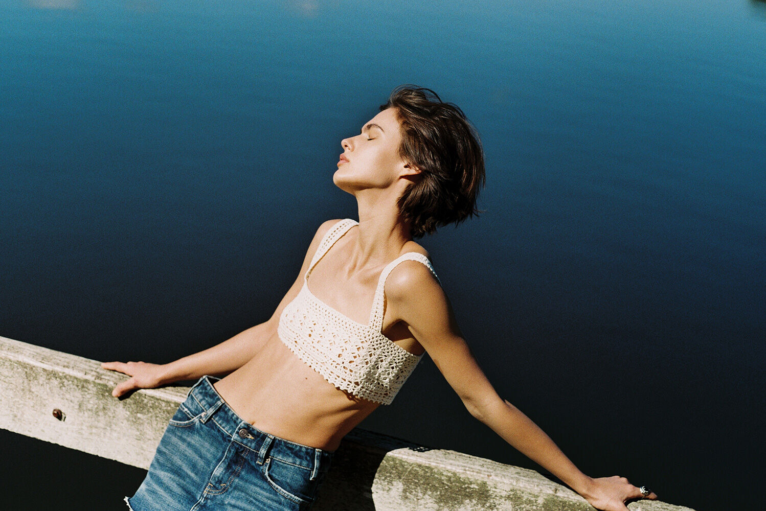 Woman in a crochet top and denim skirt leans back against a wooden rail with a blue lake and green trees blurred in the background.