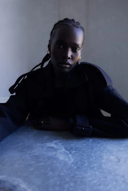 Close-up portrait of an African American woman with braided hair, wearing a black top, resting her arms on a blue marble surface.