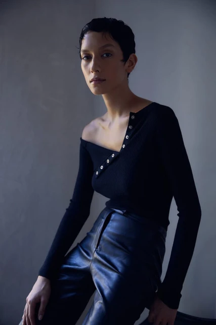 A woman with a short pixie haircut poses in a black ribbed top with leather pants against a neutral studio backdrop in a fashion setting.