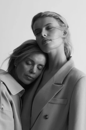 Two women with closed eyes posing closely together in tailored jackets in a monochrome portrait.