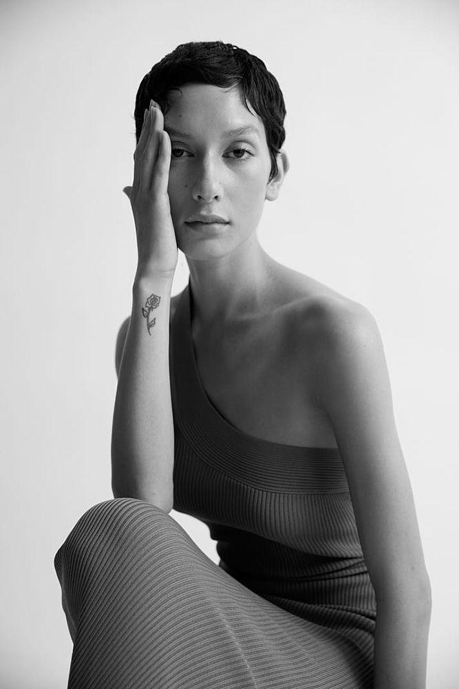 Black and white portrait of a woman with a hand on her face, short hair, and a dress, posing against a plain white backdrop.