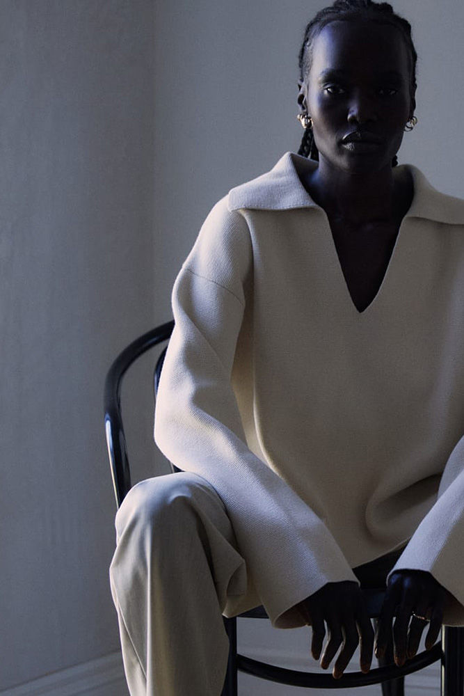 A dark-skinned woman with braids sits in a chair wearing a cream-colored, wide-collared top and matching pants in a dimly lit room.