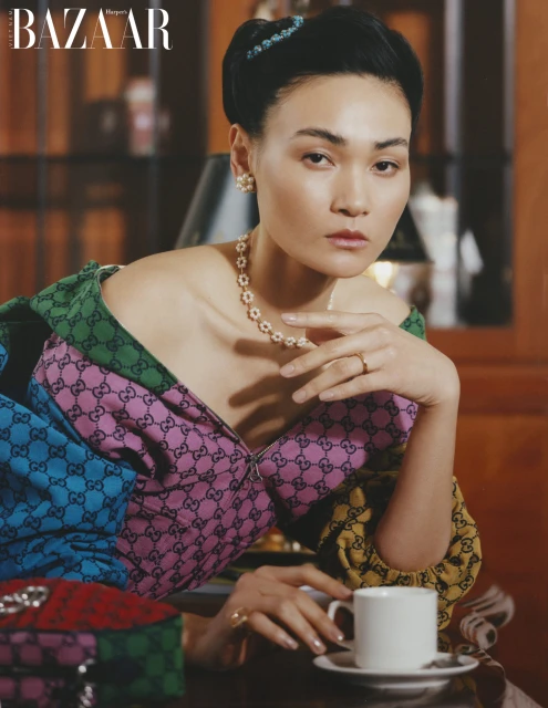 Asian woman in colorful patterned dress with pearl jewelry sits at a dark table with a teacup in a fashion editorial shot.