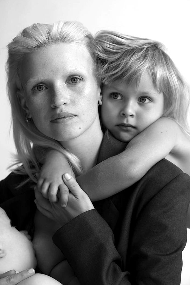 Black-and-white portrait of a blond woman holding a child wearing a blazer with a baby at her breast in a bright, indoor setting.