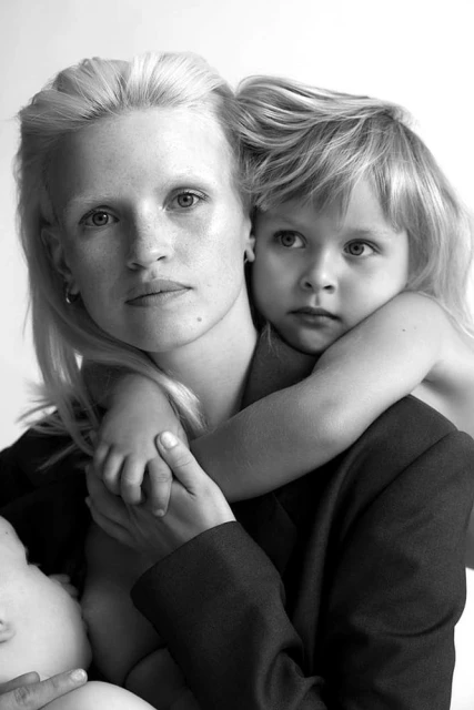 Black-and-white portrait of a blond woman holding a child wearing a blazer with a baby at her breast in a bright, indoor setting.