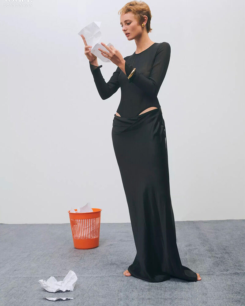 A young adult woman stands in a studio setting, wearing a long black dress, examining crumpled paper near an orange trash can.