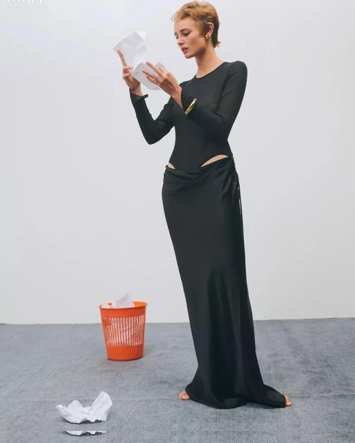 A young adult woman stands in a studio setting, wearing a long black dress, examining crumpled paper near an orange trash can.