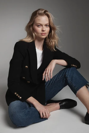 Model seated on grey floor wearing black double-breasted blazer, white t-shirt, and blue denim jeans with dark loafers.