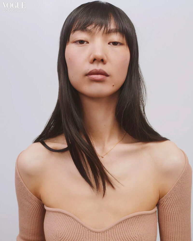 Headshot of an Asian woman with long black hair and bangs, wearing a beige sweater with off-the-shoulder sleeves, against a white background.
