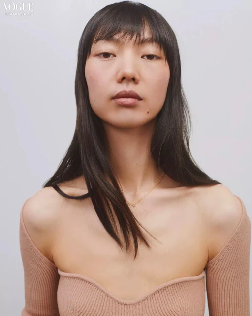 Headshot of an Asian woman with long black hair and bangs, wearing a beige sweater with off-the-shoulder sleeves, against a white background.