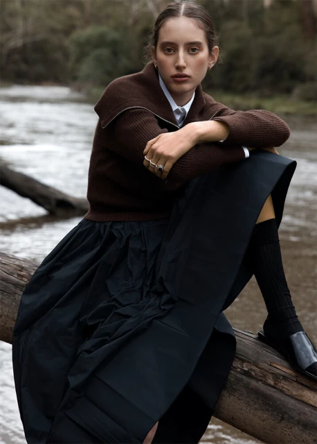 Woman in brown sweater and black skirt poses outdoors on log in a fashion editorial. River and trees in background. 
