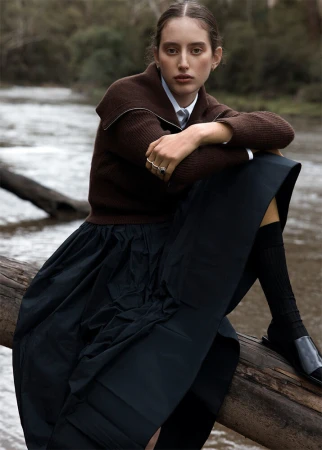 Woman in brown sweater and black skirt poses outdoors on log in a fashion editorial. River and trees in background. 