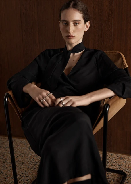 Elegant woman in a black dress sits in a modern chair in front of a wooden wall; jewelry details. Fashion editorial portrait.