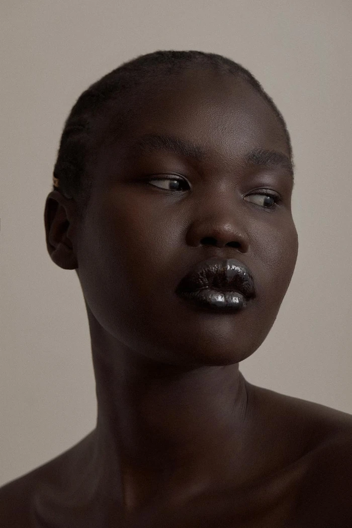 Close-up portrait of a Black woman with glossy metallic dark lips and braided hair against a neutral background