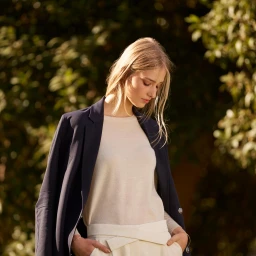 Blonde woman wearing a cream top and dark blazer outdoors with her hands in her pockets