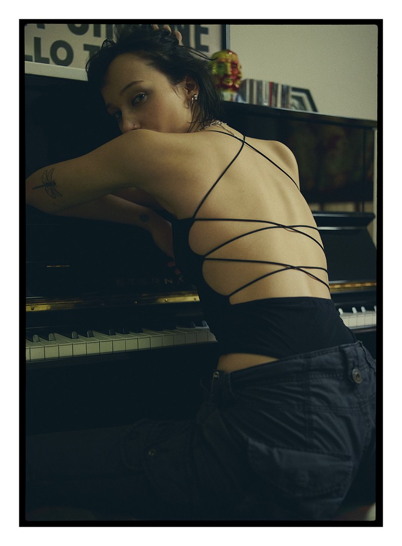 Dark-haired woman in a black top and dark shorts leans on a piano, looking at the camera in an indoor setting with art behind.