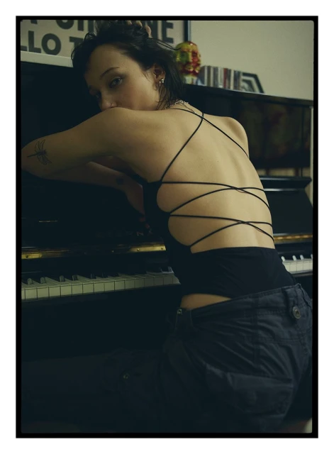 Dark-haired woman in a black top and dark shorts leans on a piano, looking at the camera in an indoor setting with art behind.