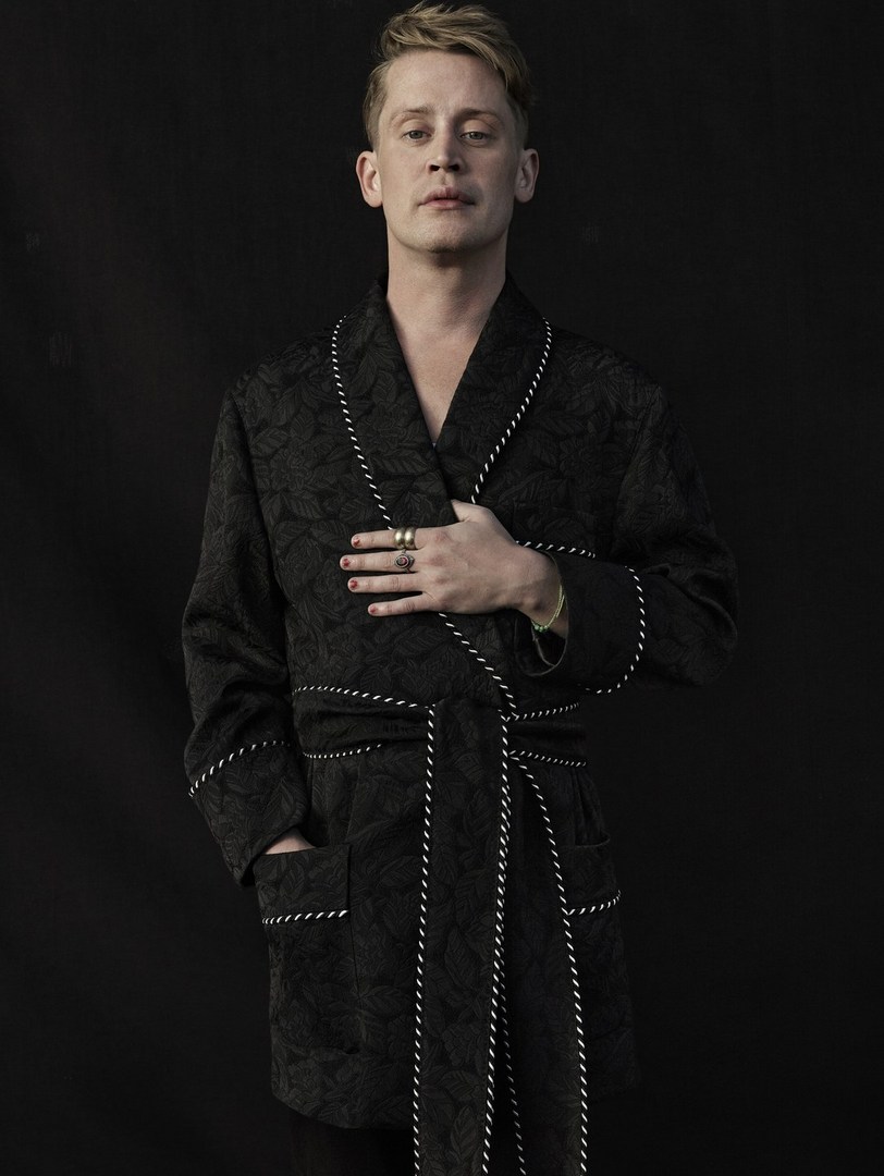 Portrait of adult male with fair skin wearing black floral robe with white piping in a photo studio against a dark grey background.