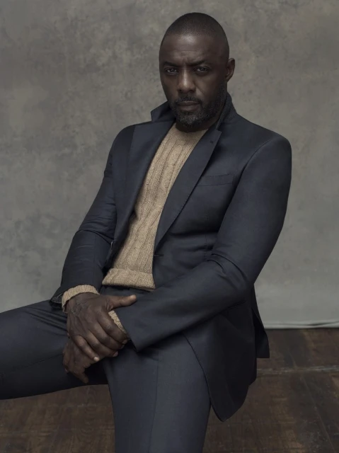 Idris Elba poses in a suit and sweater on a wooden floor. He is looking at the camera. The background is a grey wall.