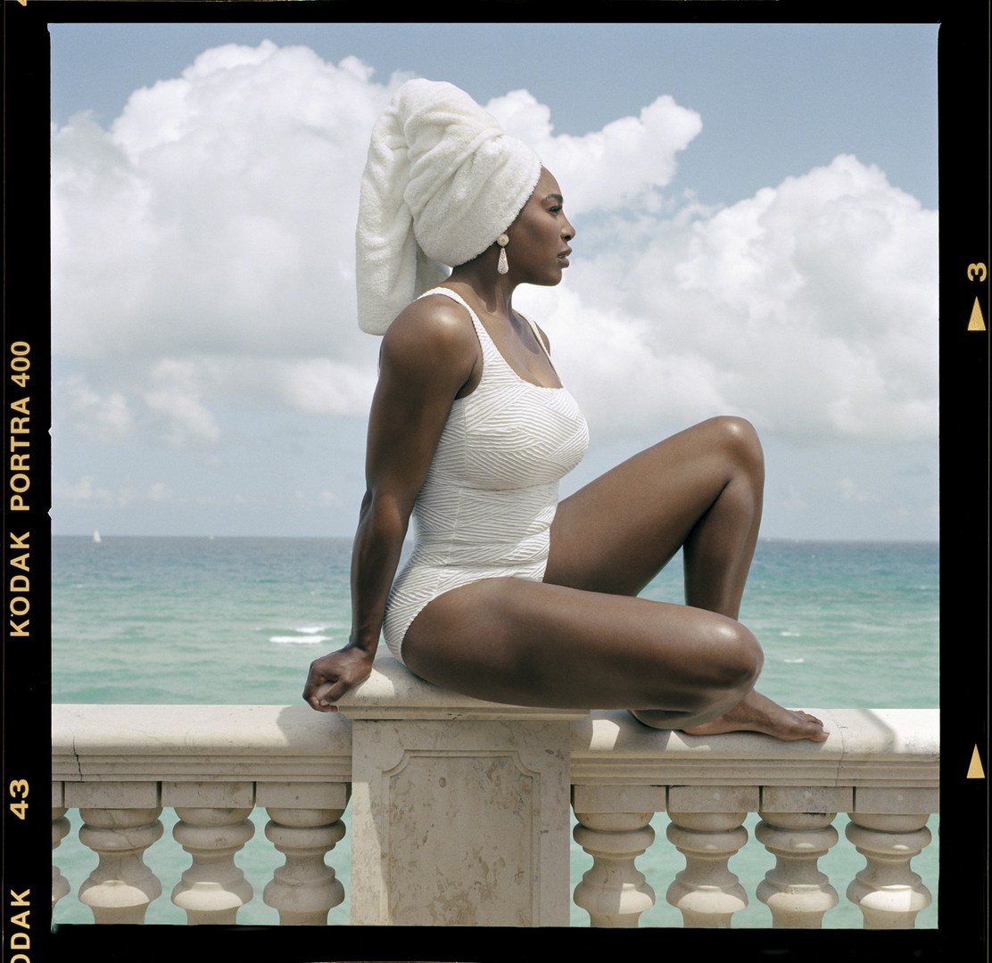 A woman in a white swimsuit and towel on her head sits on a stone railing with the ocean and sky with clouds in the background.