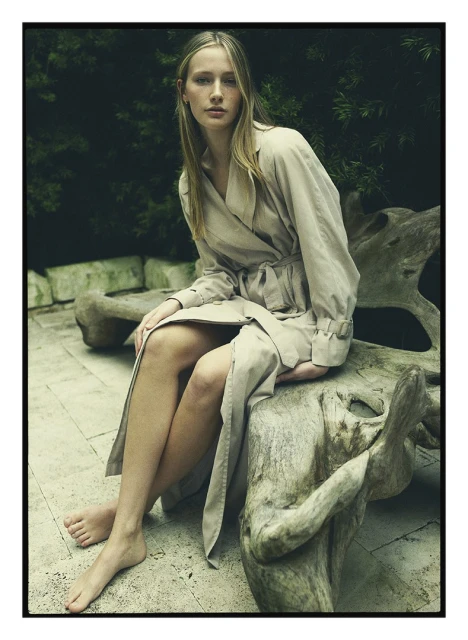 Woman in trench coat seated on a log outdoors with lush green background; a fashion shot emphasizing neutral tones and natural setting.