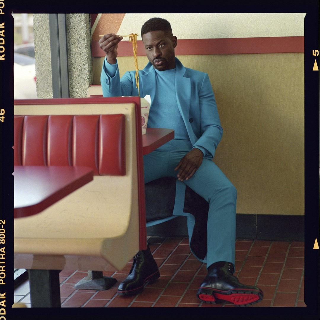 An African American man in a turquoise suit sits at a red and cream booth eating noodles with chopsticks inside a retro diner.