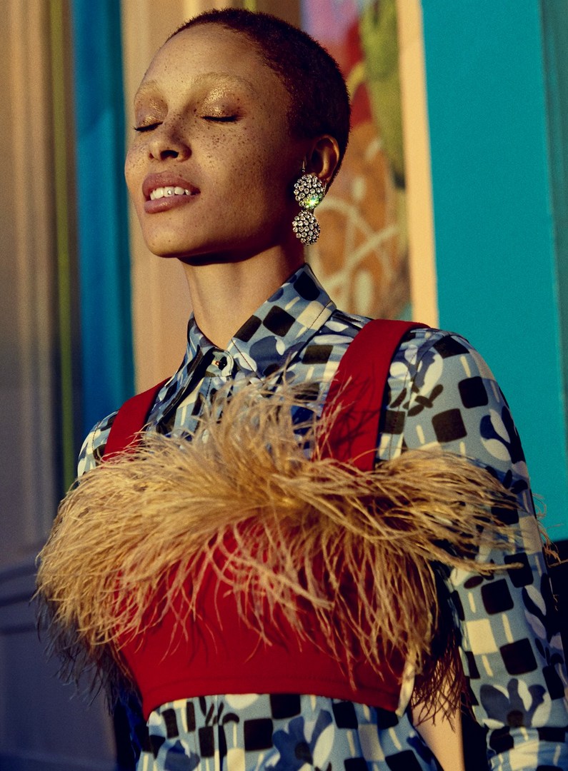 A woman with shaved head wearing a patterned top, red top, and floral earrings, poses with her eyes closed in bright sunlight.