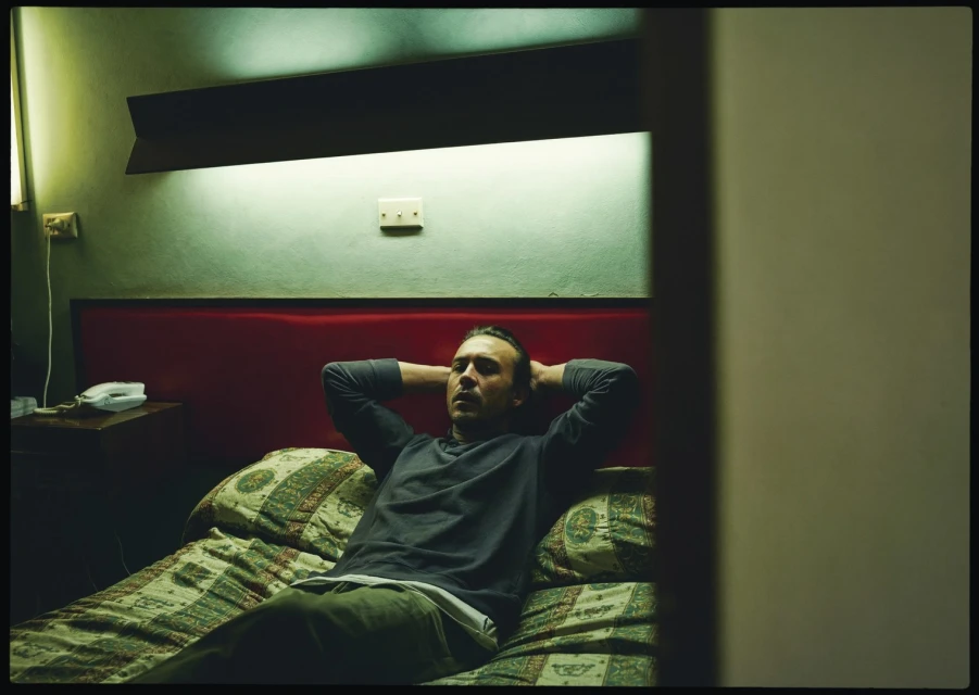 Adult man lies in bed with hands behind head in dimly lit bedroom with red headboard, patterned bedding, and vintage decor.