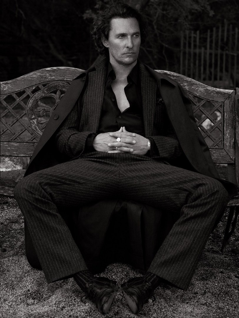 Monochrome portrait of actor Matthew McConaughey sitting on a bench in a pinstripe suit and coat, with a dark, blurred background.