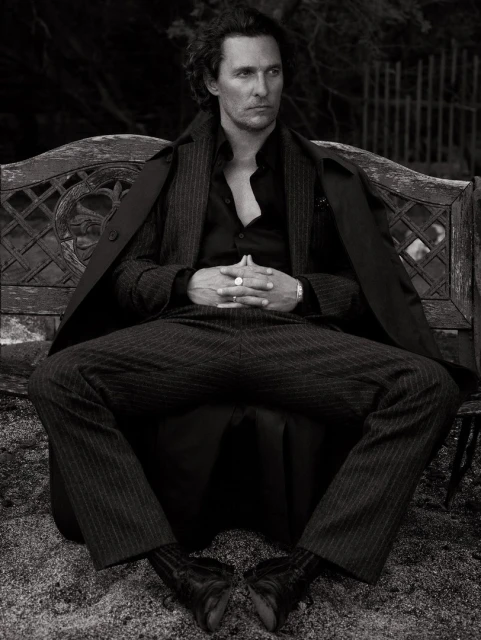 Monochrome portrait of actor Matthew McConaughey sitting on a bench in a pinstripe suit and coat, with a dark, blurred background.