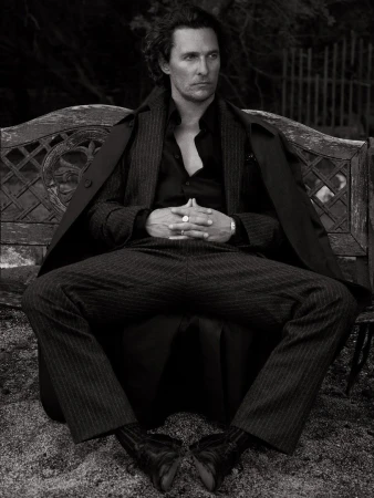 Monochrome portrait of actor Matthew McConaughey sitting on a bench in a pinstripe suit and coat, with a dark, blurred background.