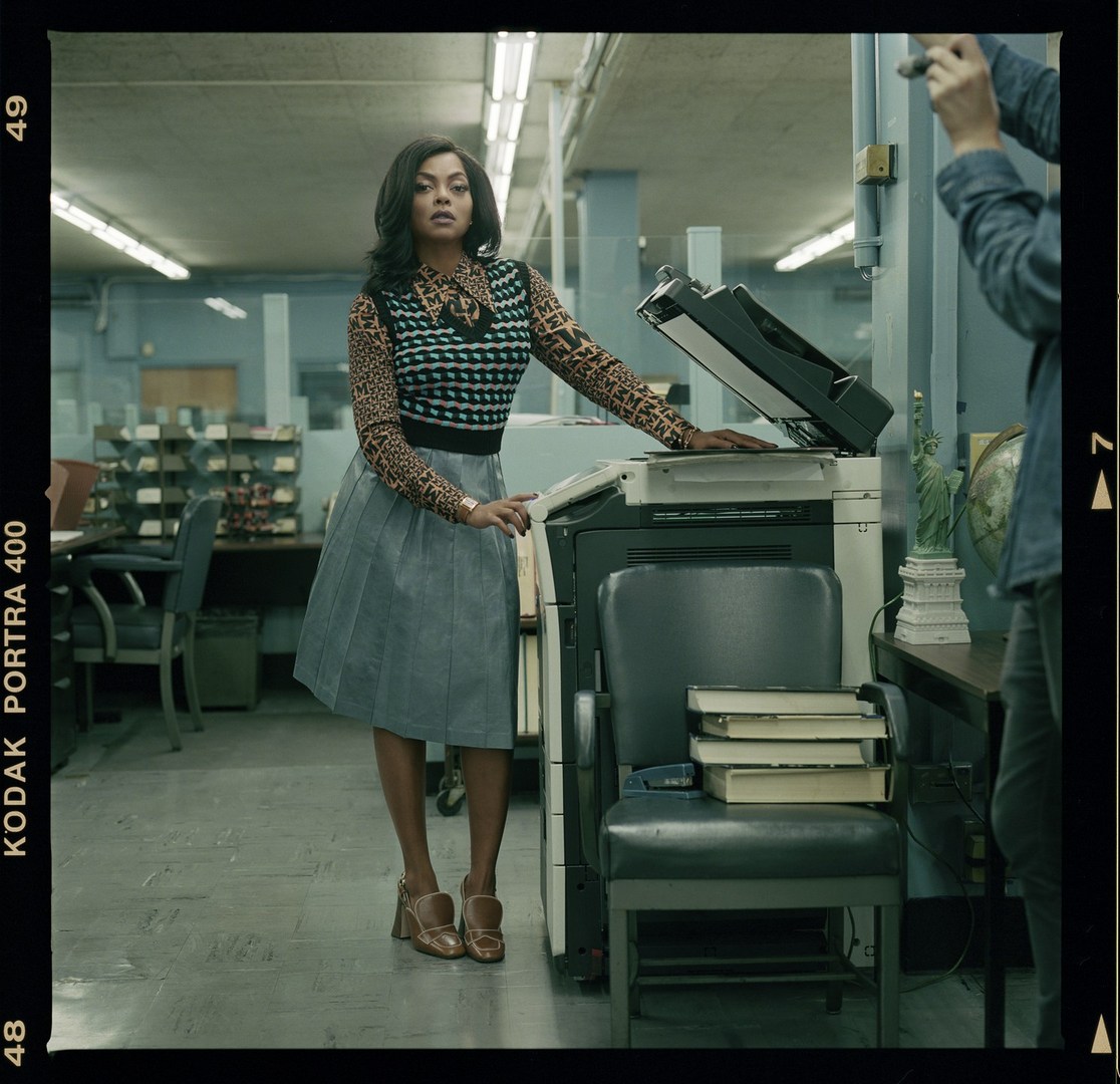 A stylish adult woman poses confidently next to an office copier in a leopard-print top and grey skirt. A photographer is visible.