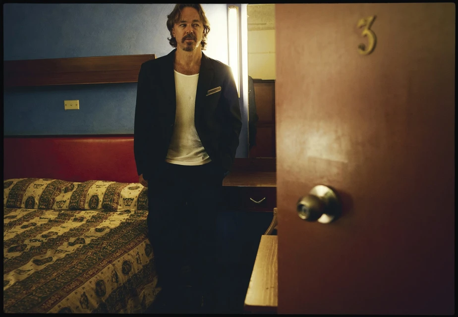 Adult male in a black suit and white shirt stands in a dimly lit motel room with a bed, peach-colored wall, and door.