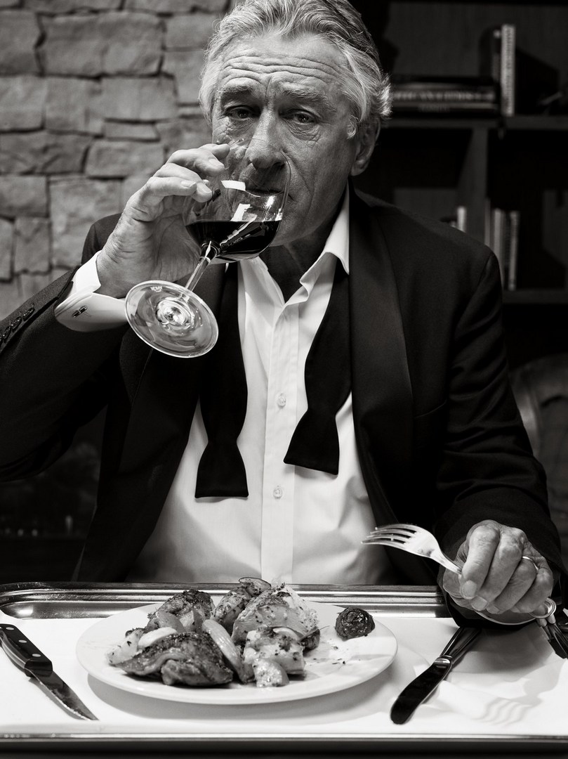 Black and white portrait of an older man in a tuxedo drinking wine with a plate of food on a tray with silverware.