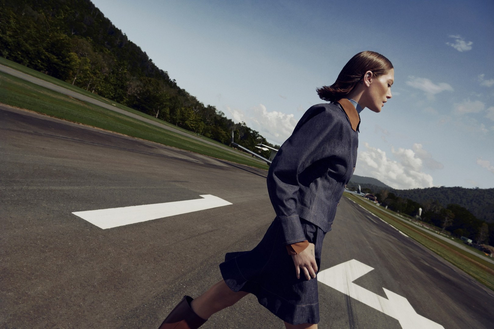 Woman in denim jacket and skirt walking on runway with mountains and sky backdrop. Fashion editorial with natural light and composition.