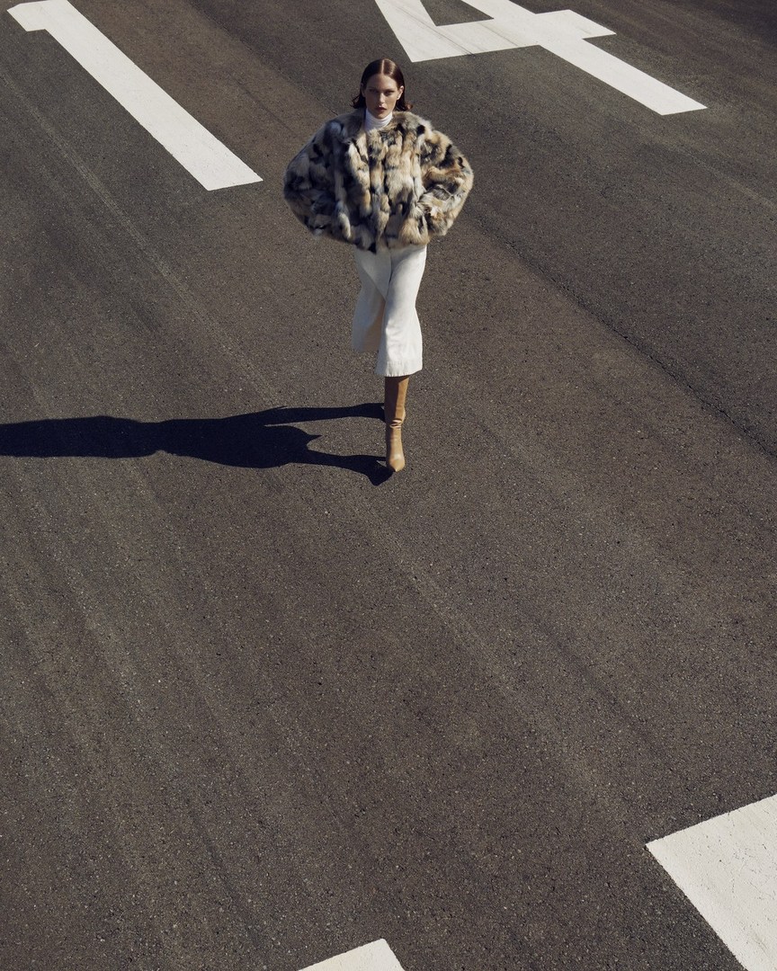 Fashion shot of an adult woman in a fur coat and boots walking on an asphalt road with white road markings in an outdoor setting.
