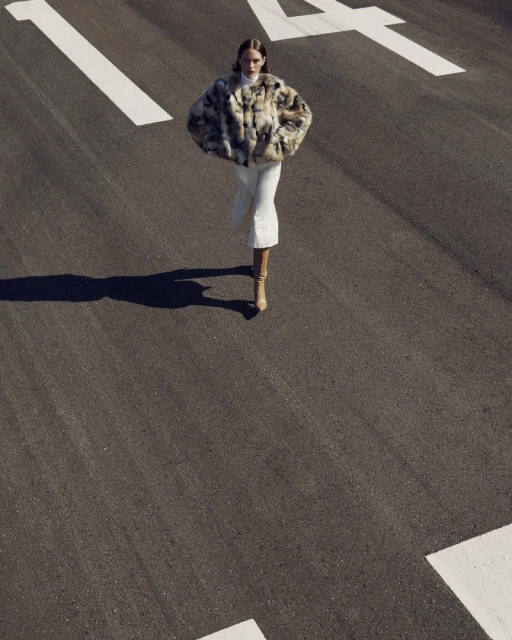 Fashion shot of an adult woman in a fur coat and boots walking on an asphalt road with white road markings in an outdoor setting.