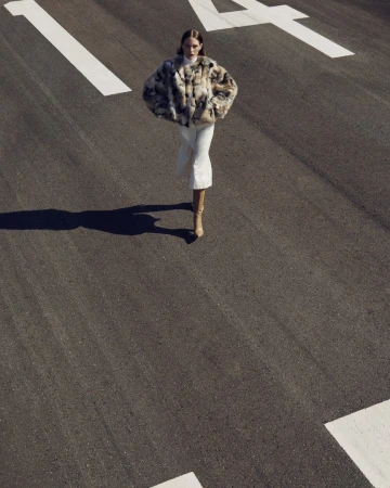 Fashion shot of an adult woman in a fur coat and boots walking on an asphalt road with white road markings in an outdoor setting.