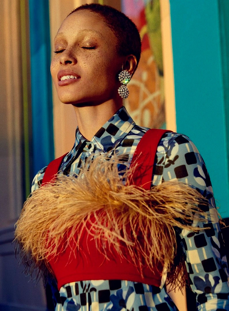 Close-up portrait of a model with short hair wearing a feathered red top over a blue patterned shirt and crystal earrings