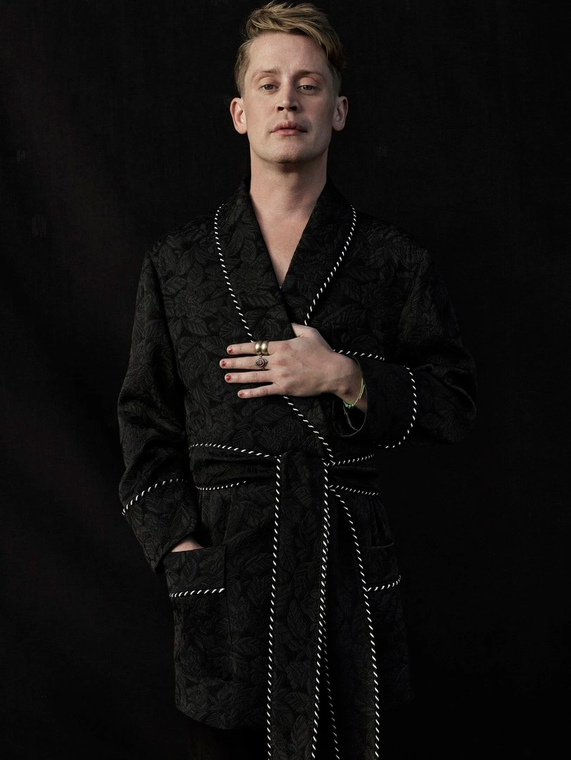Portrait of Macaulay Culkin wearing a black patterned robe with white piping against a dark background