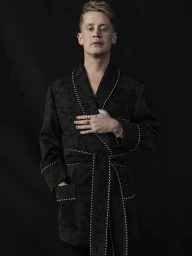 Portrait of Macaulay Culkin wearing a black patterned robe with white piping against a dark background