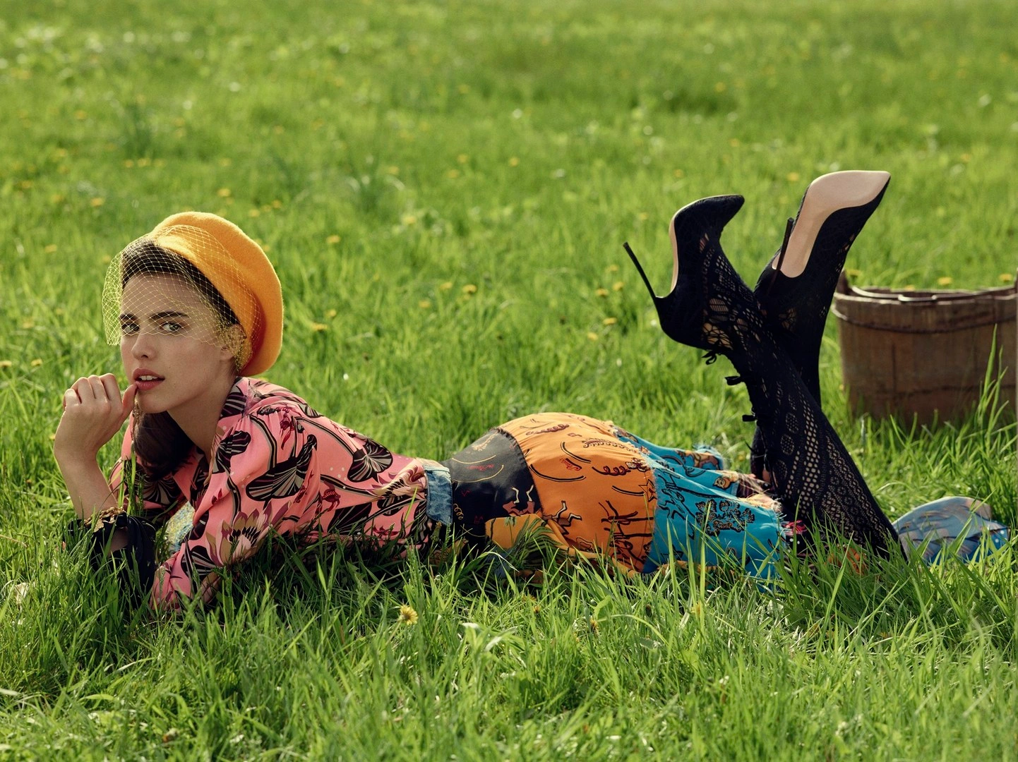 Woman in yellow beret and patterned blouse lying face down in a grassy meadow with black heels raised
