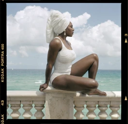 Serena Williams wearing a white one-piece swimsuit and towel wrap seated on a balcony overlooking the ocean, by Robbie Fimmano
