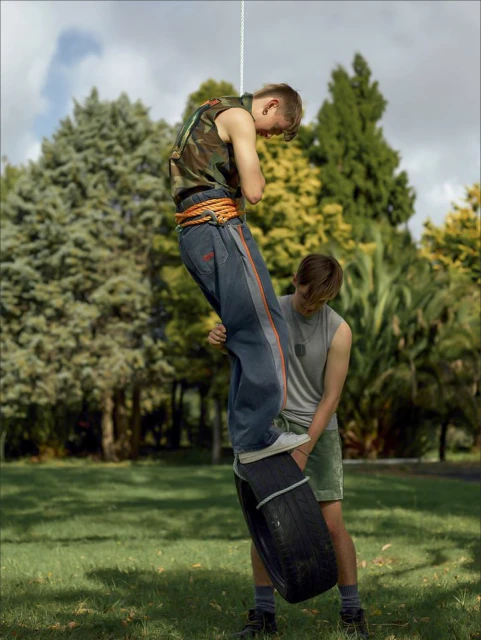 Two young men outdoors on a makeshift tire swing suspended from a rope in a grassy park with leafy trees in the background.