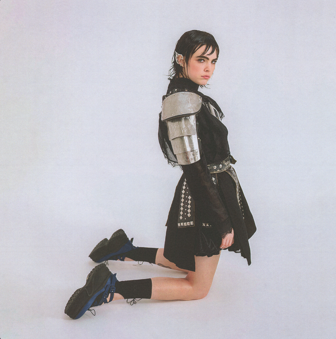 Young woman in a black outfit with silver armor pieces poses kneeling in a photo studio with a white background and studio lighting.