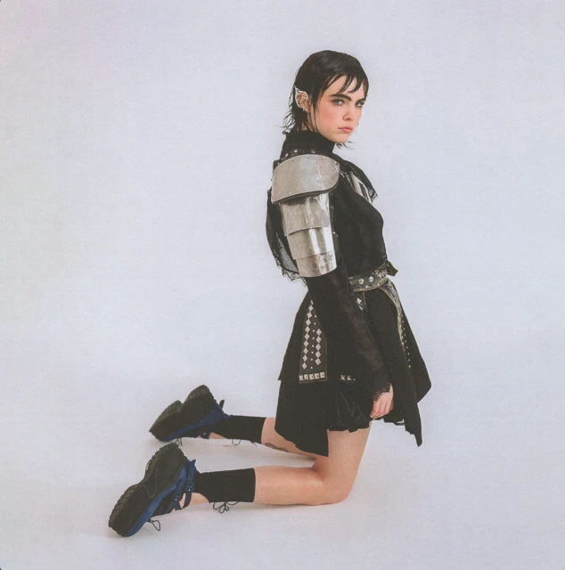 Young woman in a black outfit with silver armor pieces poses kneeling in a photo studio with a white background and studio lighting.