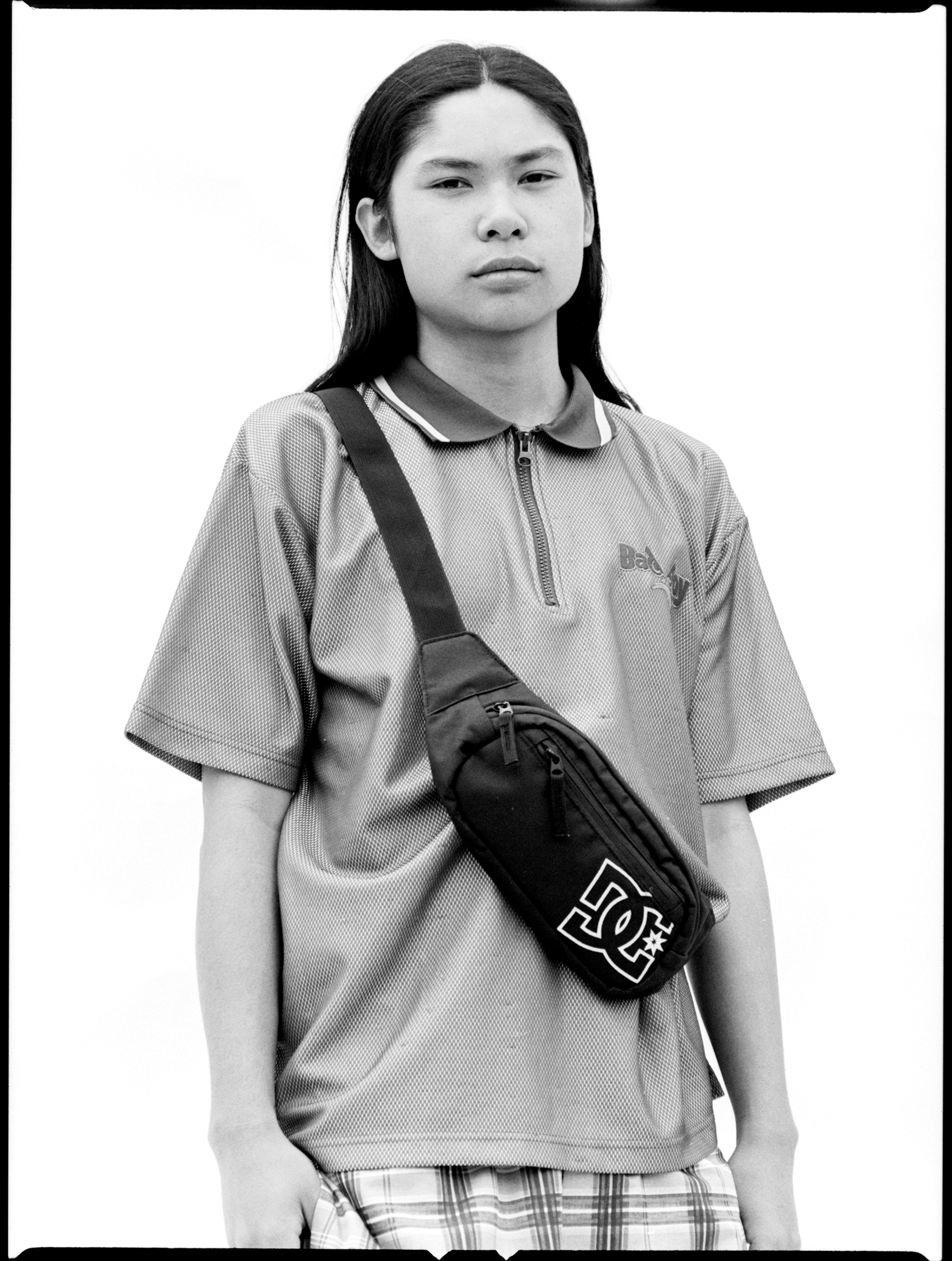 Monochrome portrait of a young person wearing a branded fanny pack and a collared shirt against a bright, neutral background.