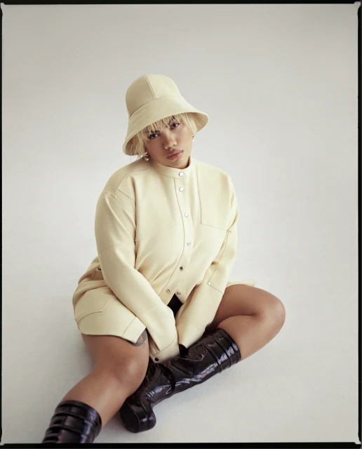 A stylish woman in an ivory jacket and bucket hat poses in a studio setting with glossy black boots, casting a calm, confident gaze.