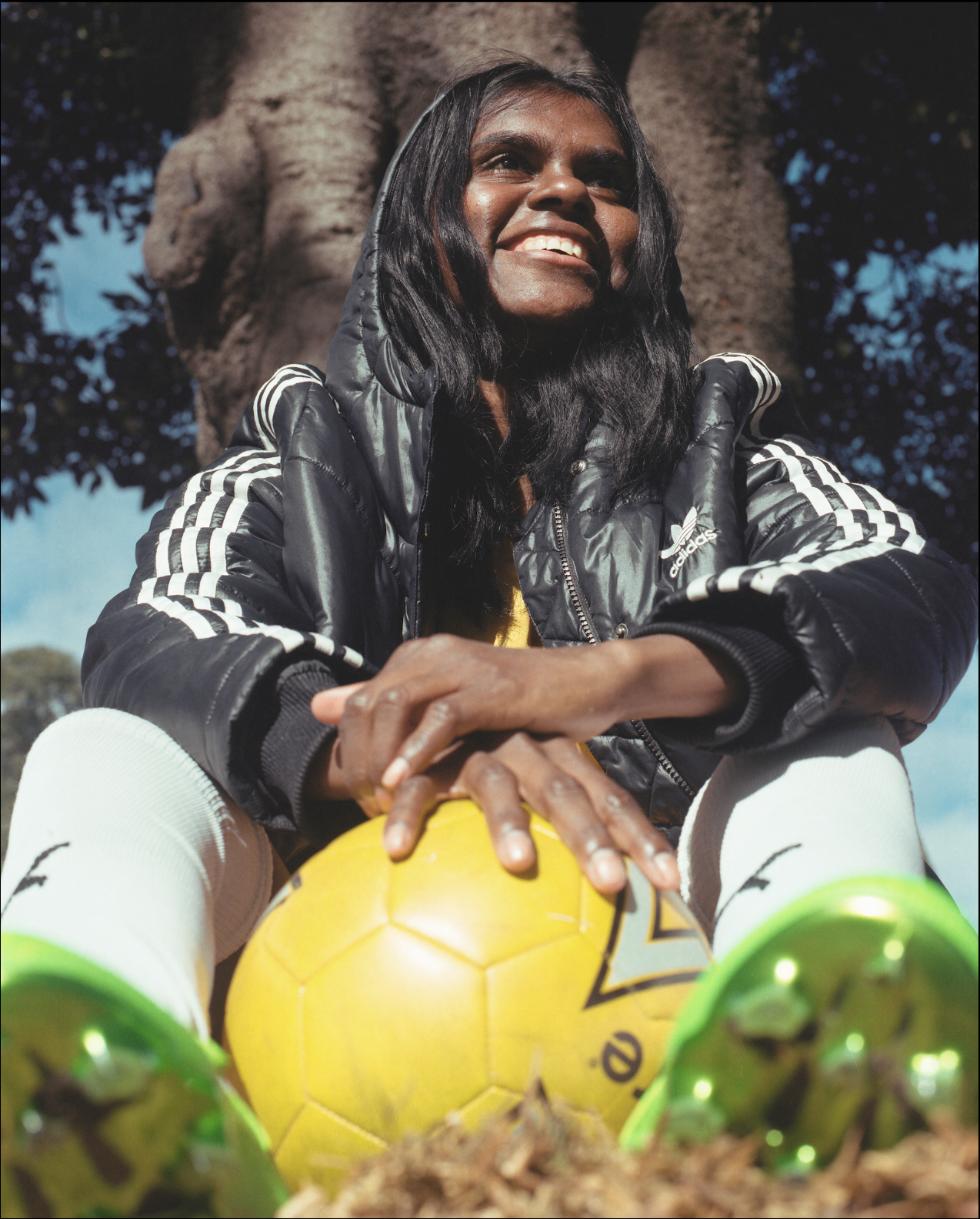 Female soccer player smiles, sits with ball and turf shoes outdoors. Wearing Adidas jacket, white socks. Tree and sky background.